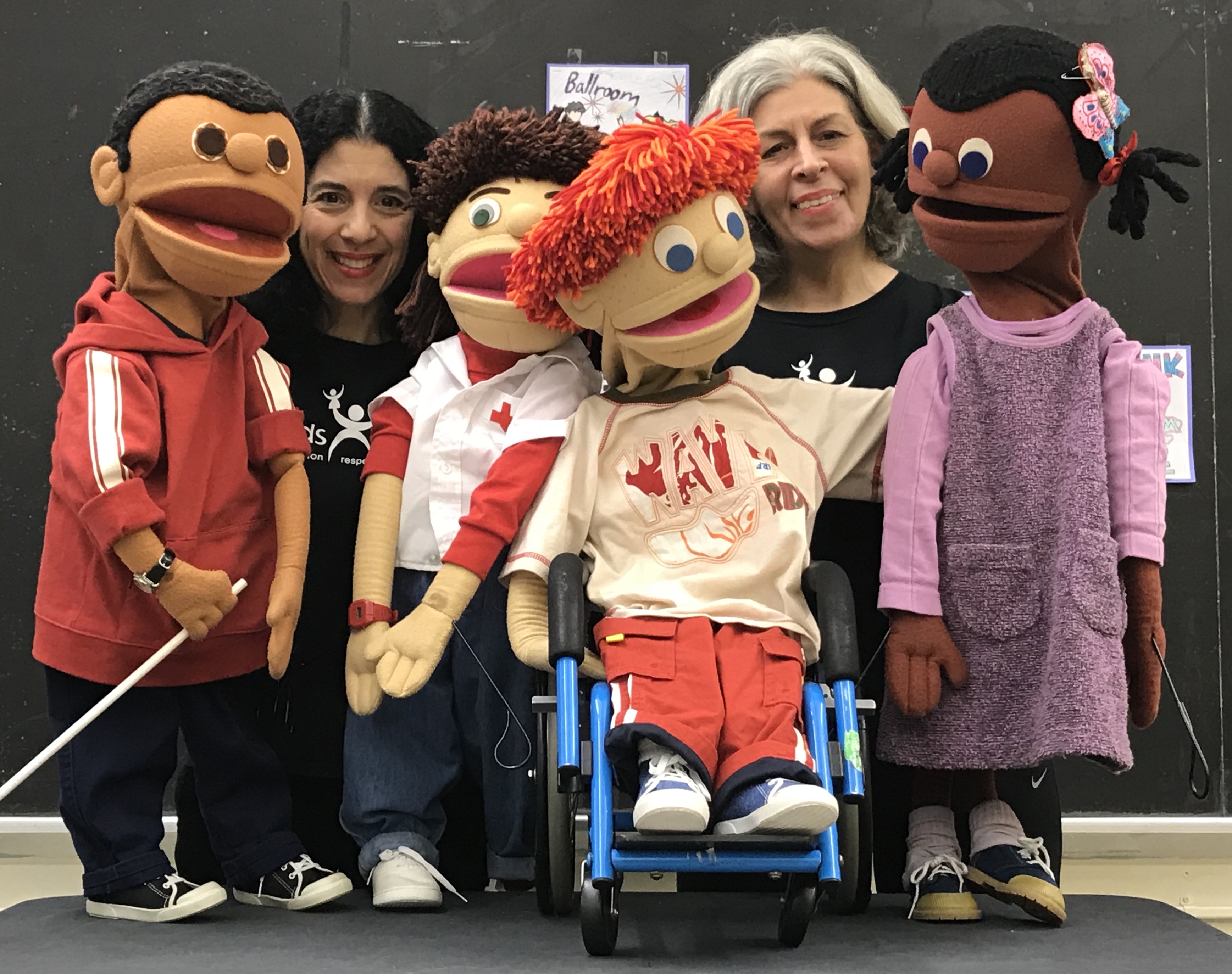 A group of two adult women and four "kid" puppets. One puppet is in a wheelchair, three are standing.